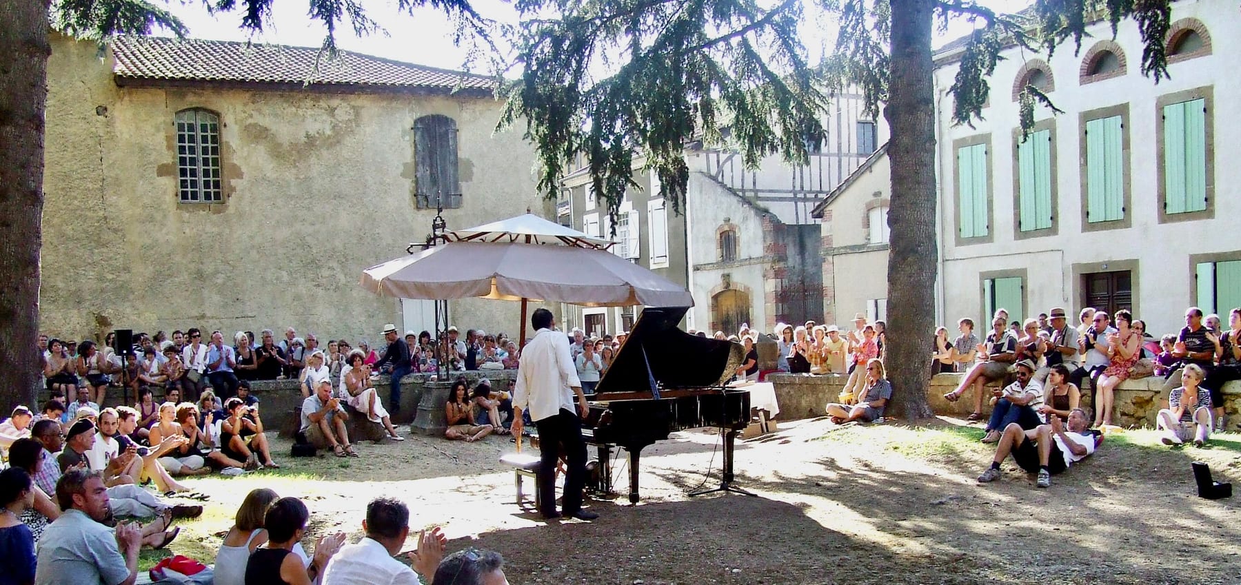 Concert de piano en plein air sous les arbres, public assis dans la cour